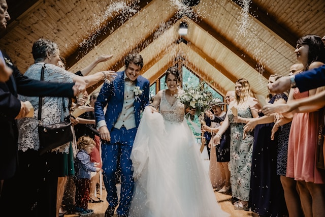 A bride and groom walking back down the aisle in a wooden room as guests throw rice over them.