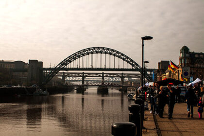 A curved bridge crosses a still river. People gather around market stalls on a quay to the right of the image.